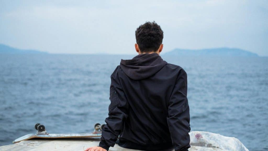 A man sits on a rock by the sea, looking out at the water on a cloudy day.