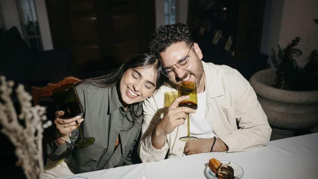 A woman leaning on a man’s shoulder as they have beer while seated at a table in a cafe.