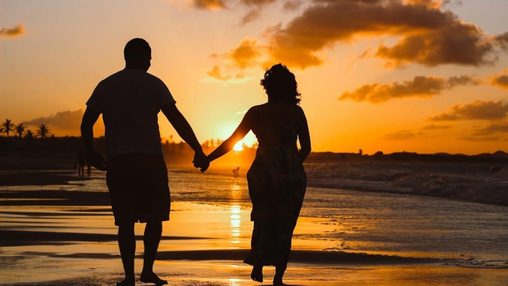 A silhouette of a couple walking while holding hands down a beach.