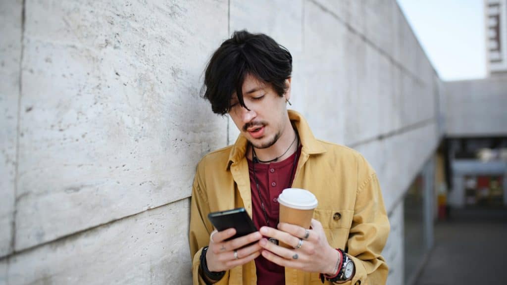 A mustached man holding a coffee mug and looking at his phone while leaning against a wall outdoors.