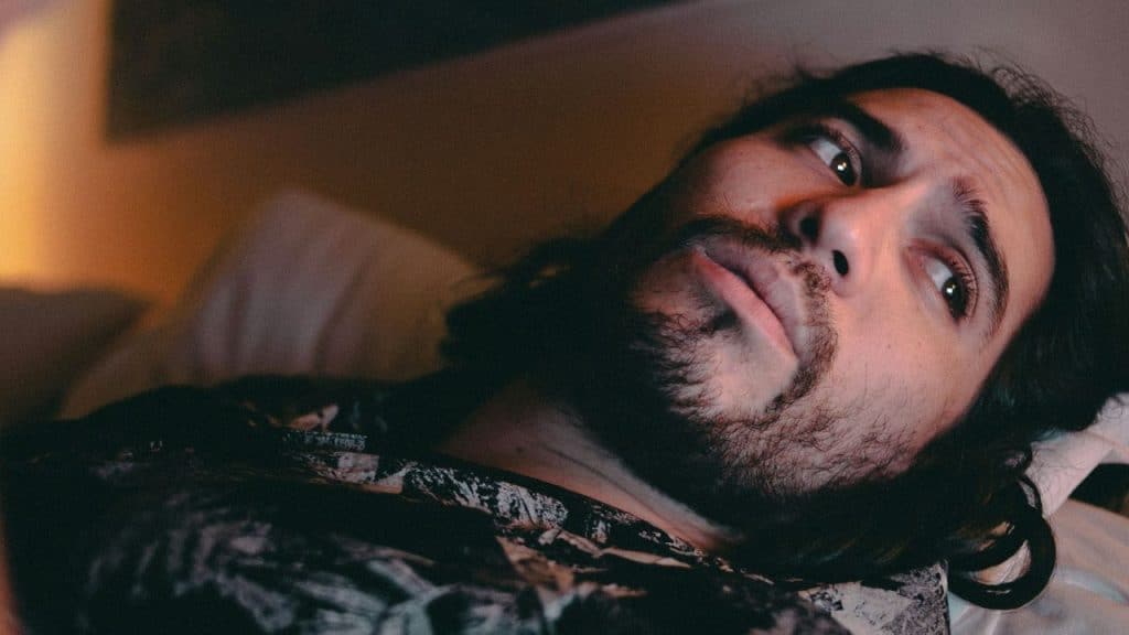 A close-up of a man with a beard looking up while lying in a dimly lit setting.