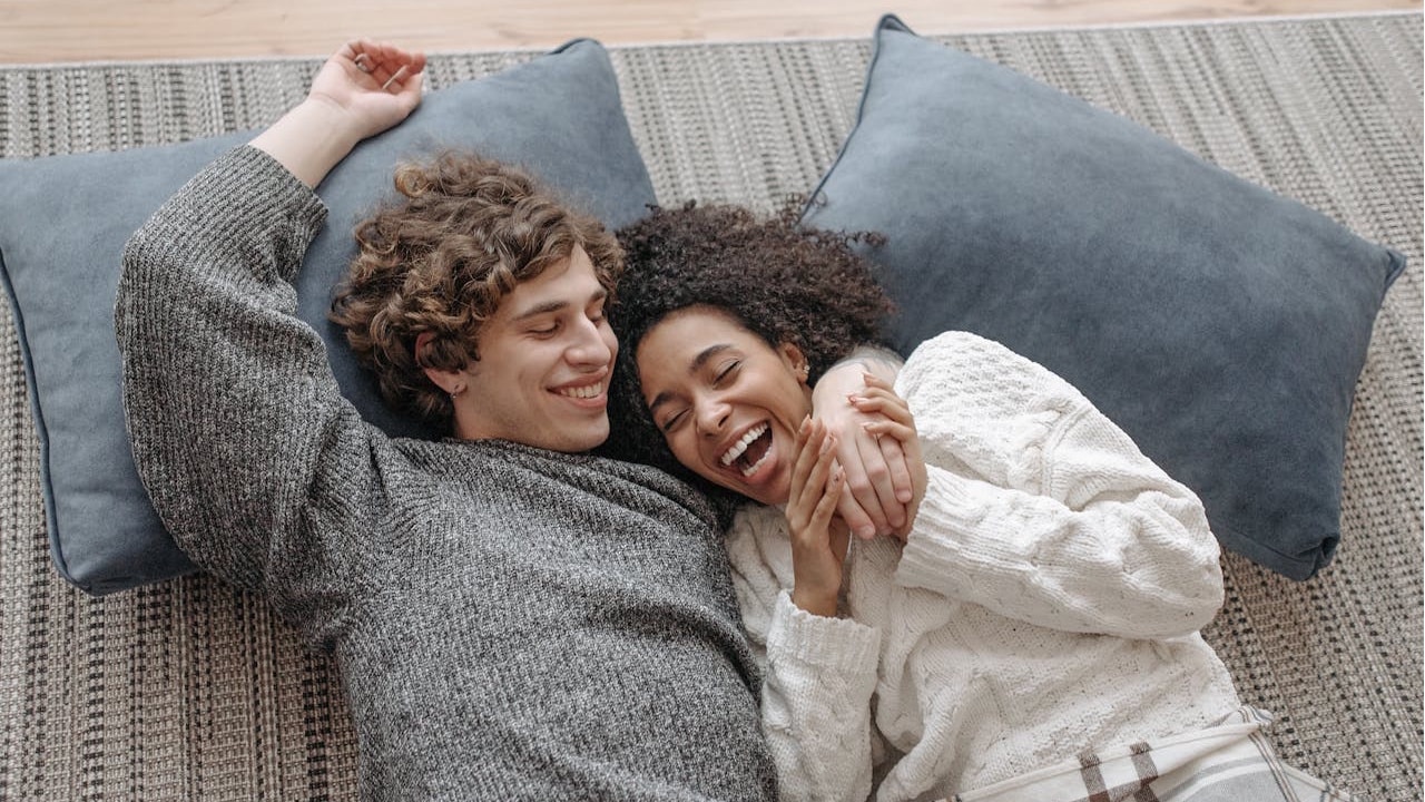 A smiling man and woman lying on a rug with pillows.