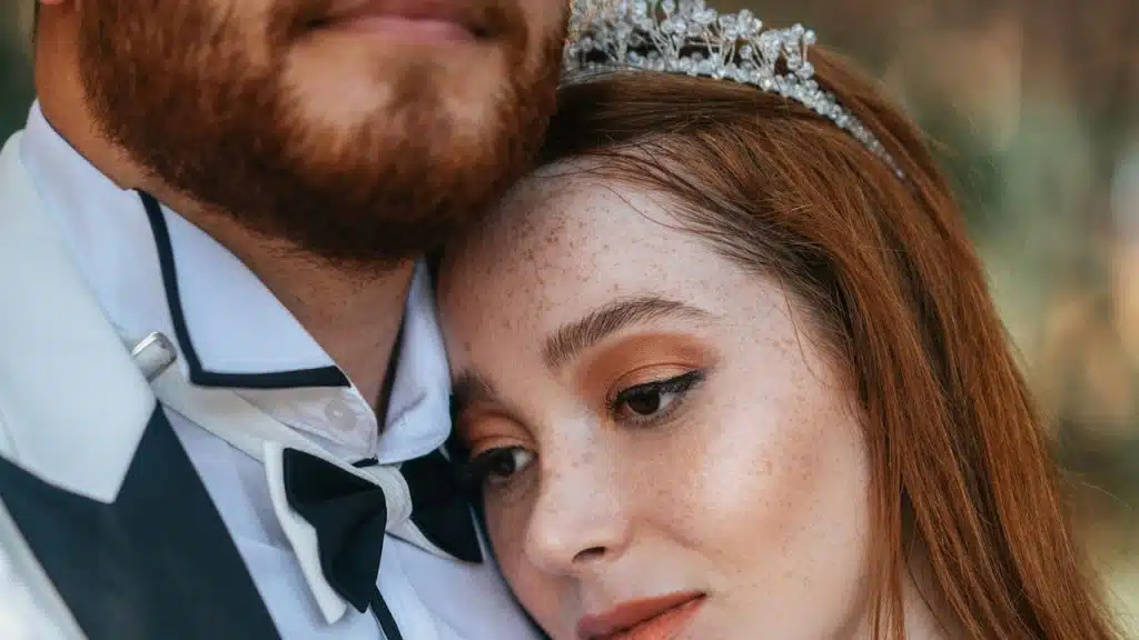 A woman wearing a tiara is resting her head on a bearded man’s chest.