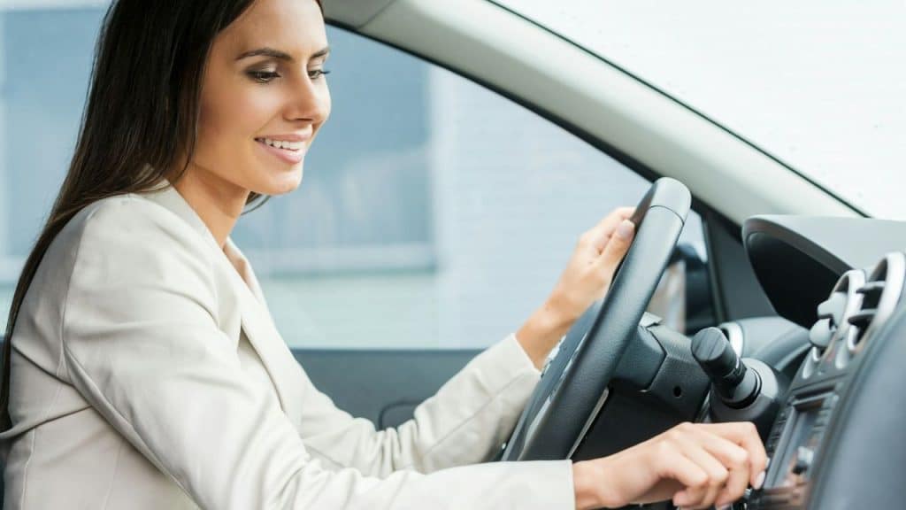 A smiling young woman in a white blazer is driving a car.