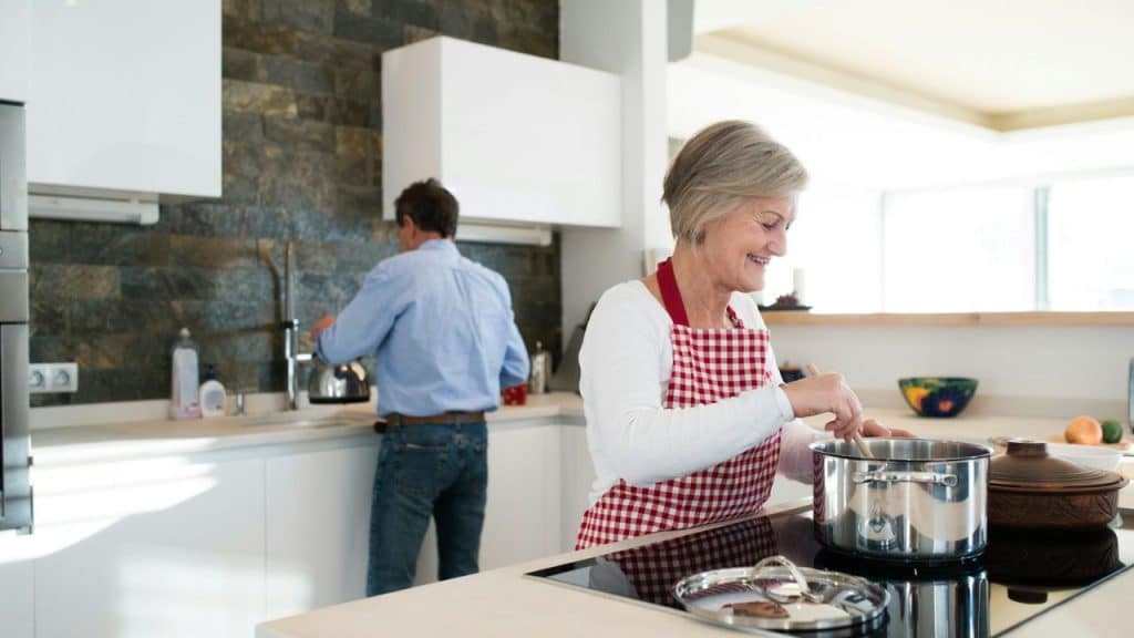 A smiling middle-aged woman is cooking with a pot in a brightly lit kitchen.