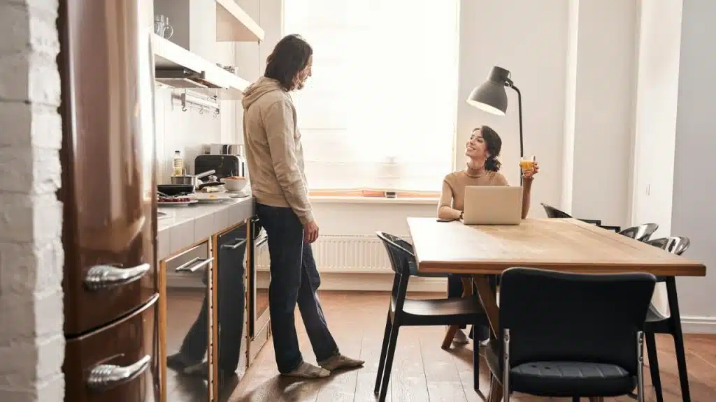Man leaning on a kitchen counter; woman with a laptop and a drink at a table.