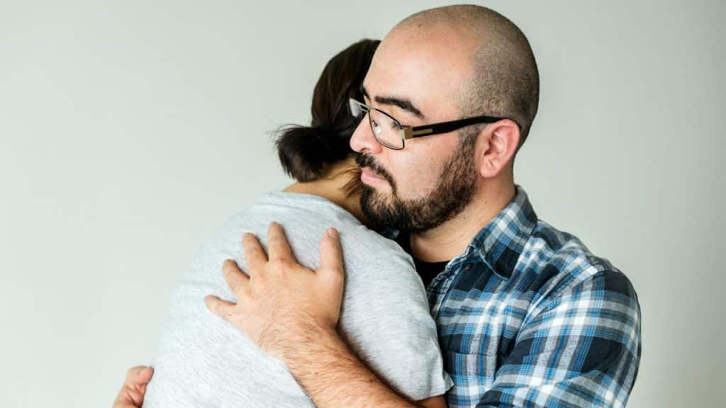 A bearded man in glasses is hugging a person with their back to the camera.