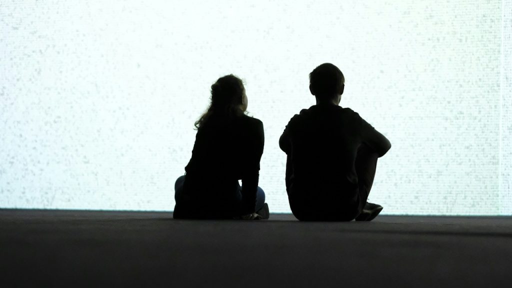 A couple sitting cross legged on the ground together with their backs to the camera.