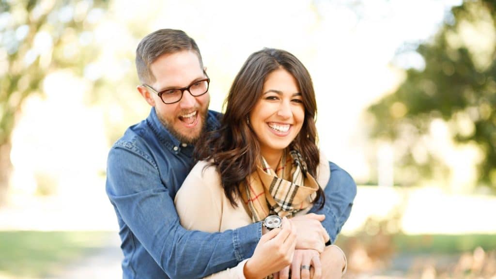 A man with glasses is hugging a smiling woman from behind in an outdoor setting.