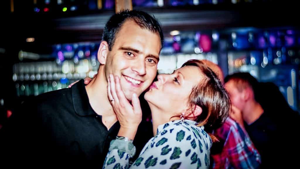 A woman kisses a smiling man's cheek in a dimly lit, crowded space, likely a bar or club.