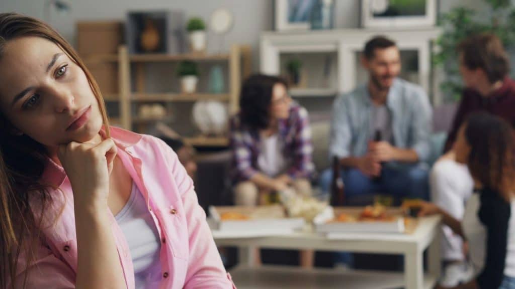 A woman with a pink shirt looks sad while friends party in the background.