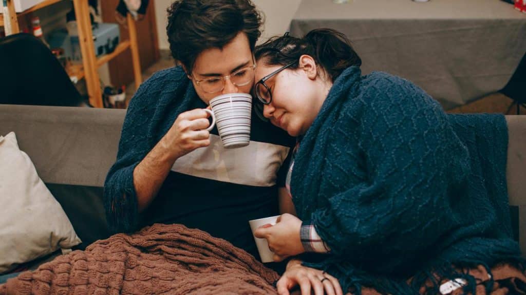 A couple cuddled under blankets while sharing warm drinks on a couch.