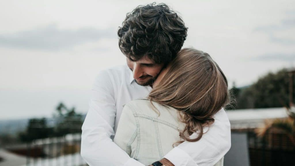 A man and woman embrace outdoors against a cloudy, soft-focus background.