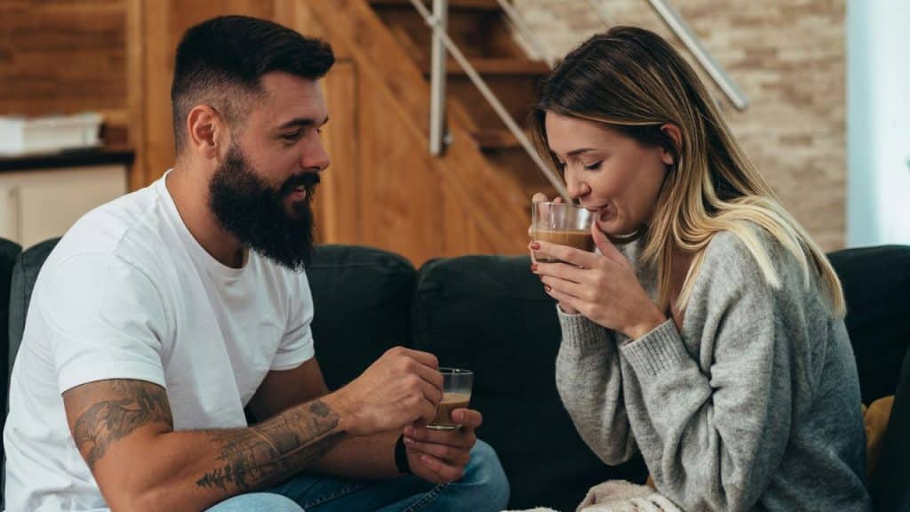 Bearded man and woman drinking coffee and looking at each other on a couch.