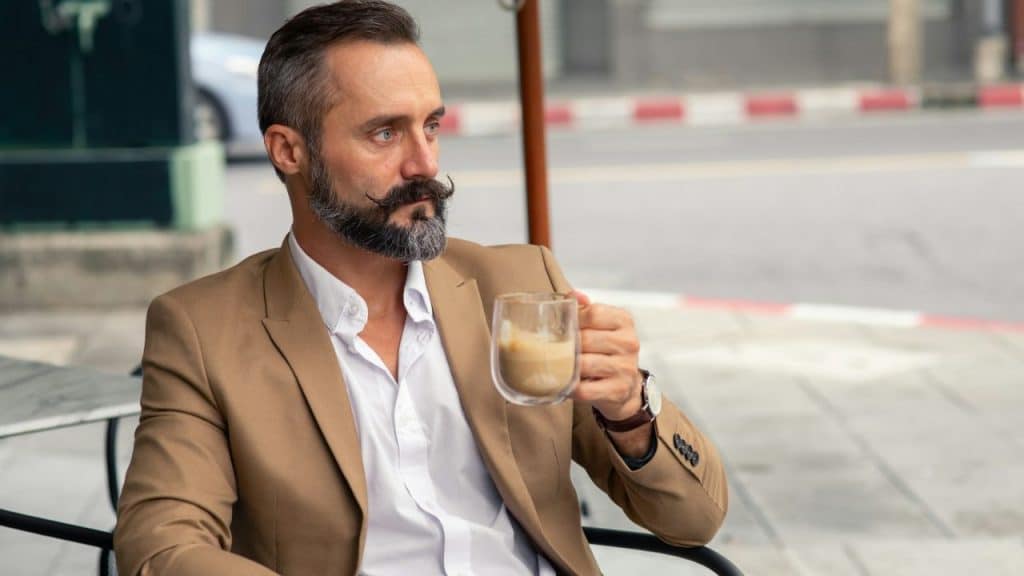 A man with a styled beard holds a glass coffee cup while sitting outdoors, looking away.