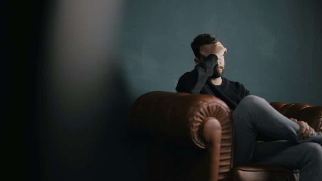 A man sits on a leather armchair, holding his hand to his forehead, looking stressed.