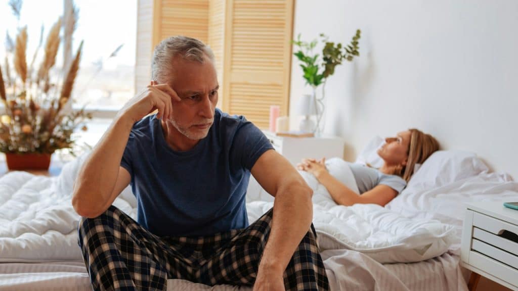 A man is sitting on the edge of the bed while his sick wife is lying on the bed.