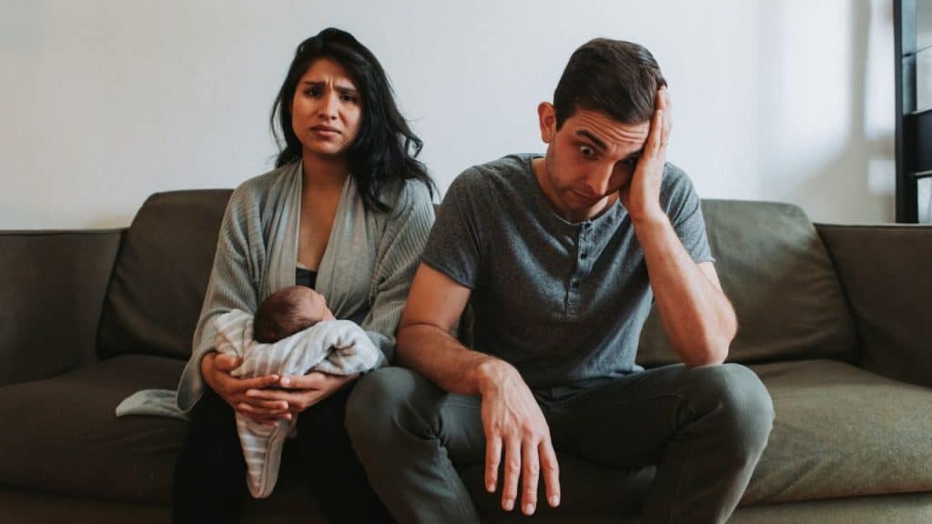 A worried couple sits on a couch; the woman is holding a newborn baby.