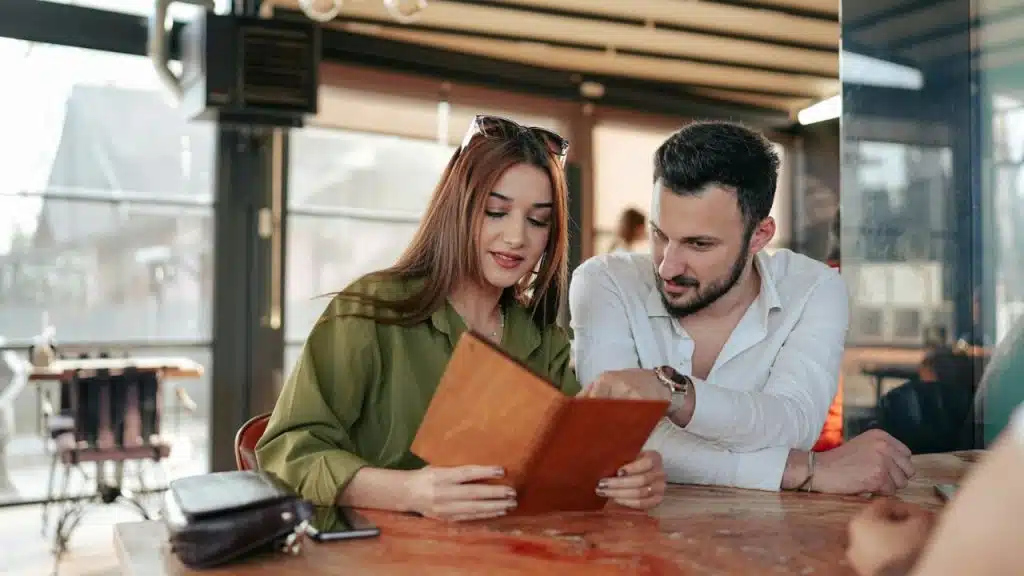 A couple sits at a table, looking at a brown menu together in a sunny restaurant.