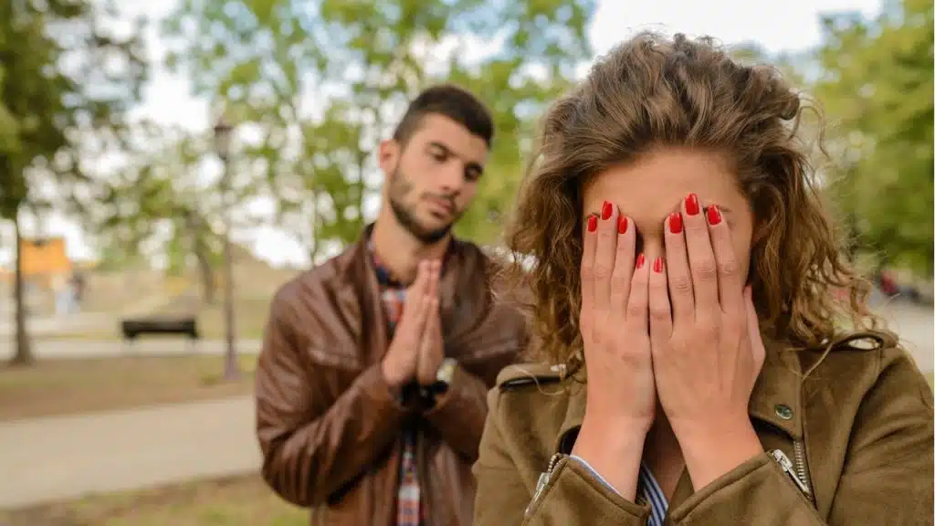 A woman in a park covers her face with her hands, while a man out of focus in the background holds his hands together as if pleading.
