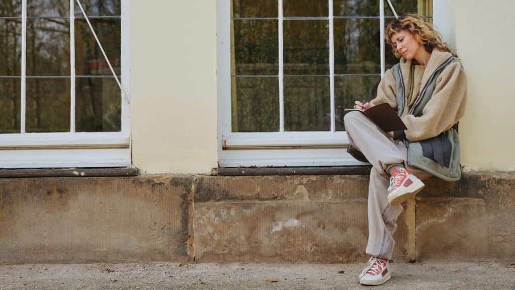 Woman sits writing in a notebook next to a large window outside a building.