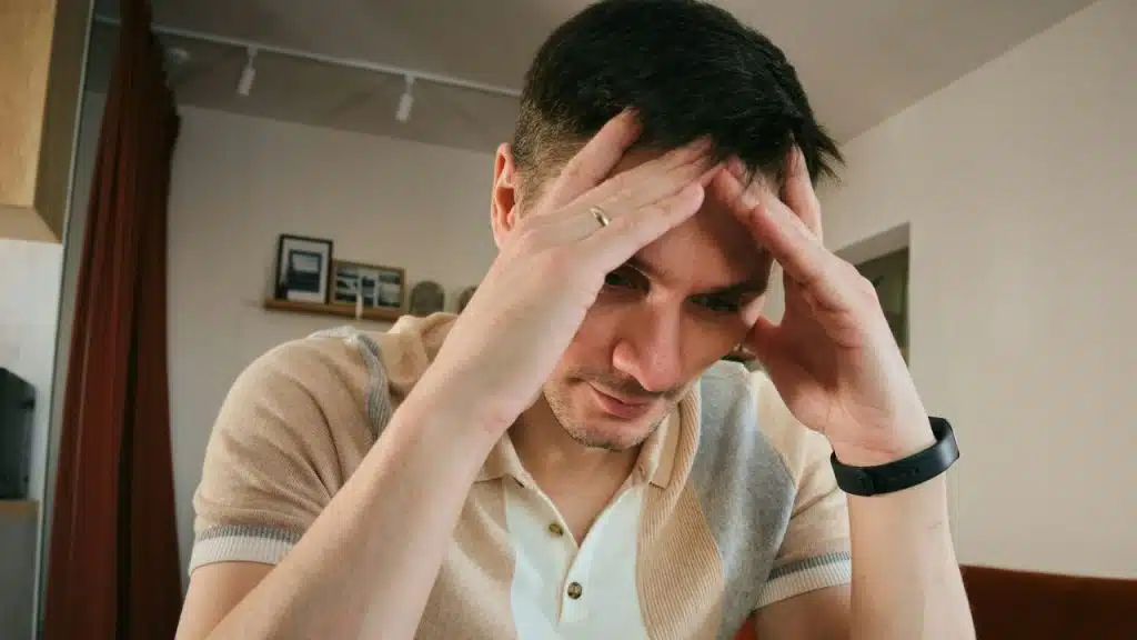 Man in a collared shirt holding his head with both hands, looking down anxiously.