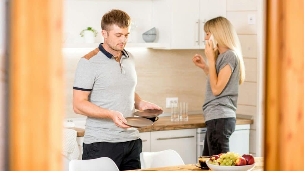 A man sets plates down while a woman talks on the phone in a modern kitchen.