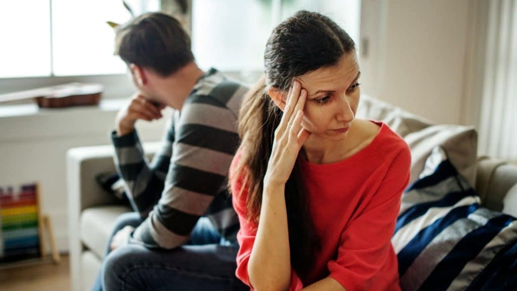 Woman in red shirt looks stressed, sitting back-to-back with a man on a couch.