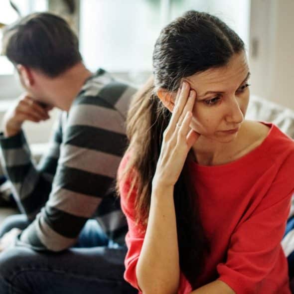 Woman in red shirt looks stressed, sitting back-to-back with a man on a couch.