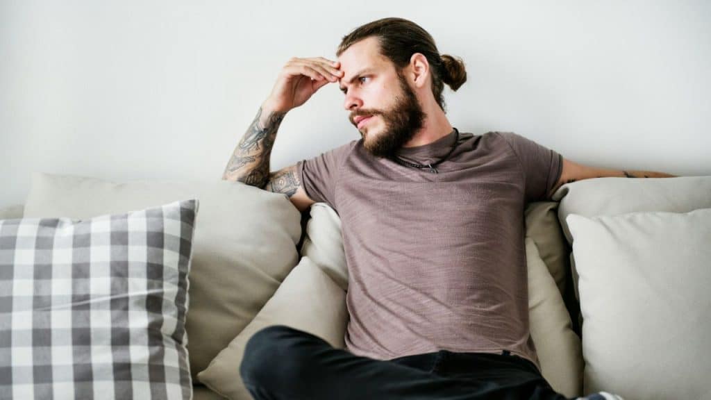 A man in a brown shirt is sitting on a sofa with his hand on his forehead.