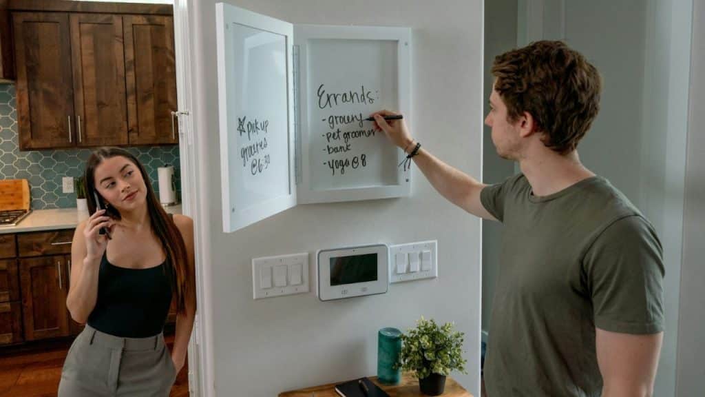 A man writes on a whiteboard while a woman talks on a phone in a kitchen.