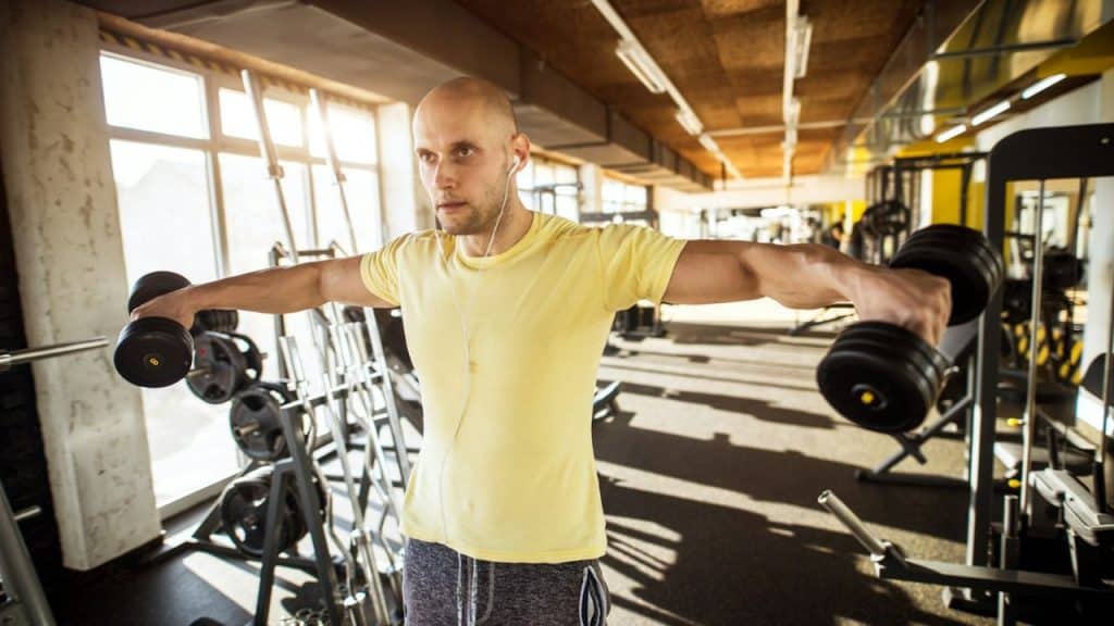 A bald man with earphones is doing lateral raises with black dumbbells in a gym.