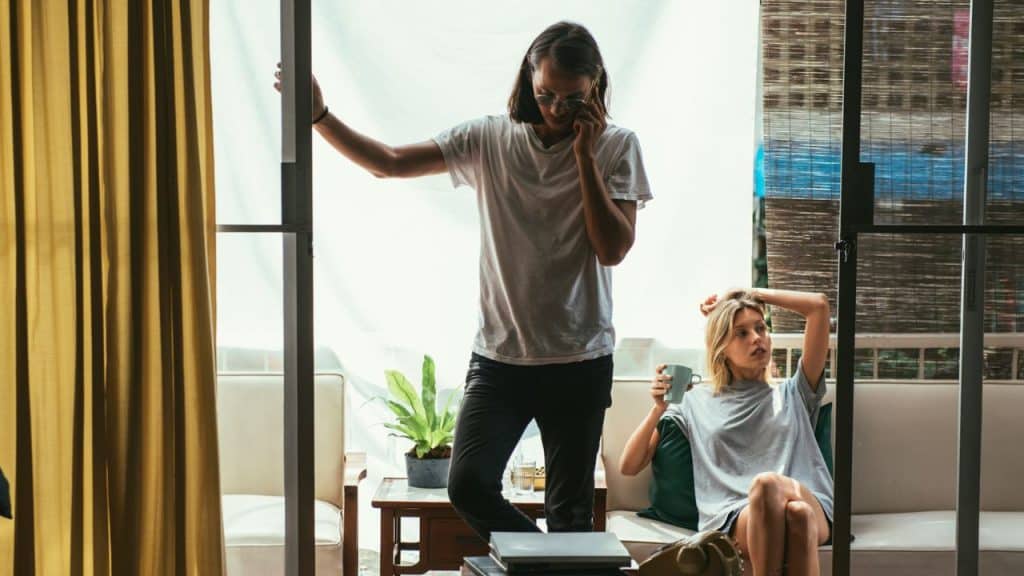 A man on the phone stands by a door while a woman sits on a couch holding a cup.