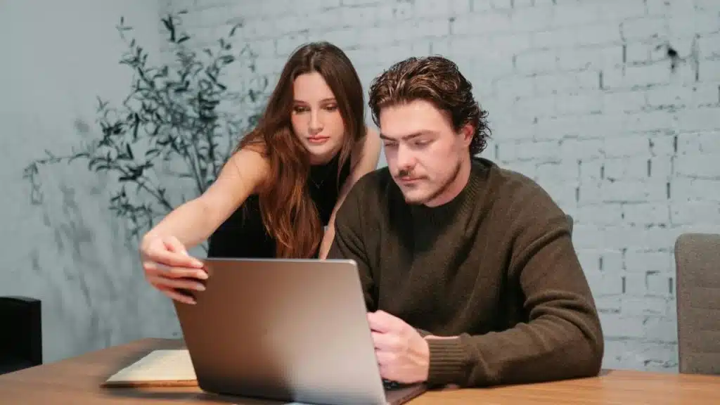 A man and woman lean over a laptop on a desk, intently focused on the screen.