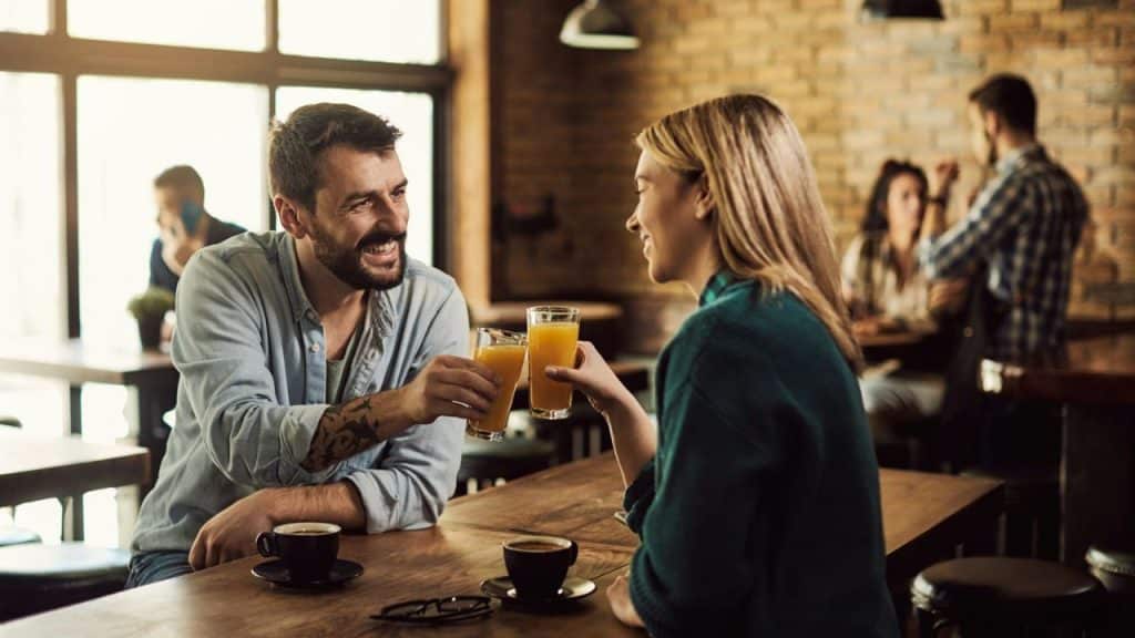 Man and woman smiling and toasting orange juice in a dimly lit cafe.