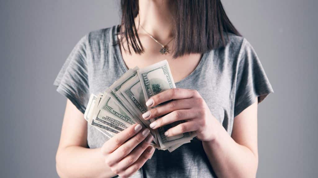 A woman in a gray shirt holding a fan of US dollar bills.
