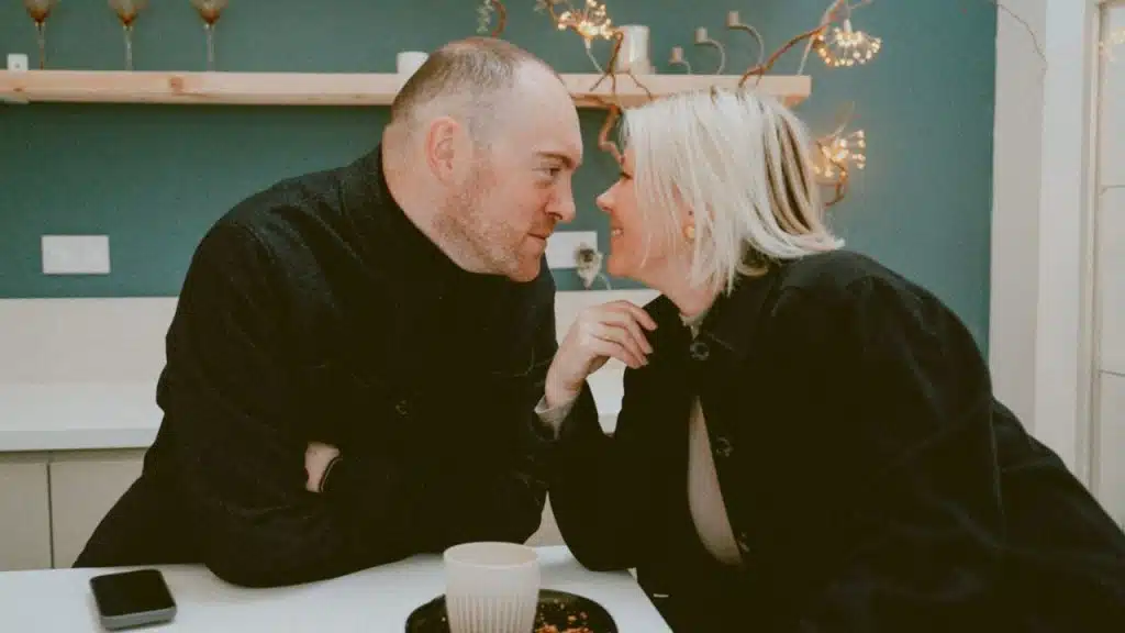 Man and woman, both in dark jackets, lean in toward each other, smiling, across a table.