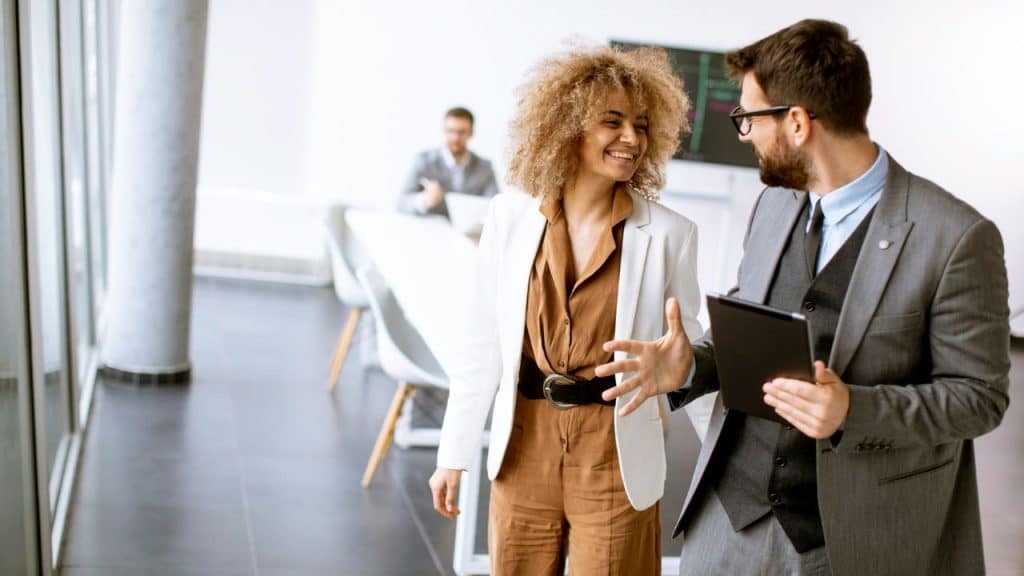 A man and a woman in business attire are smiling and talking in a bright office.