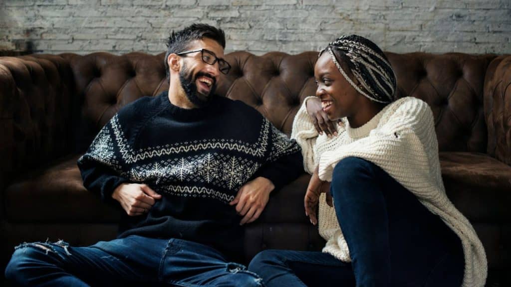 A smiling man and woman sit on the floor in front of a brown leather couch.