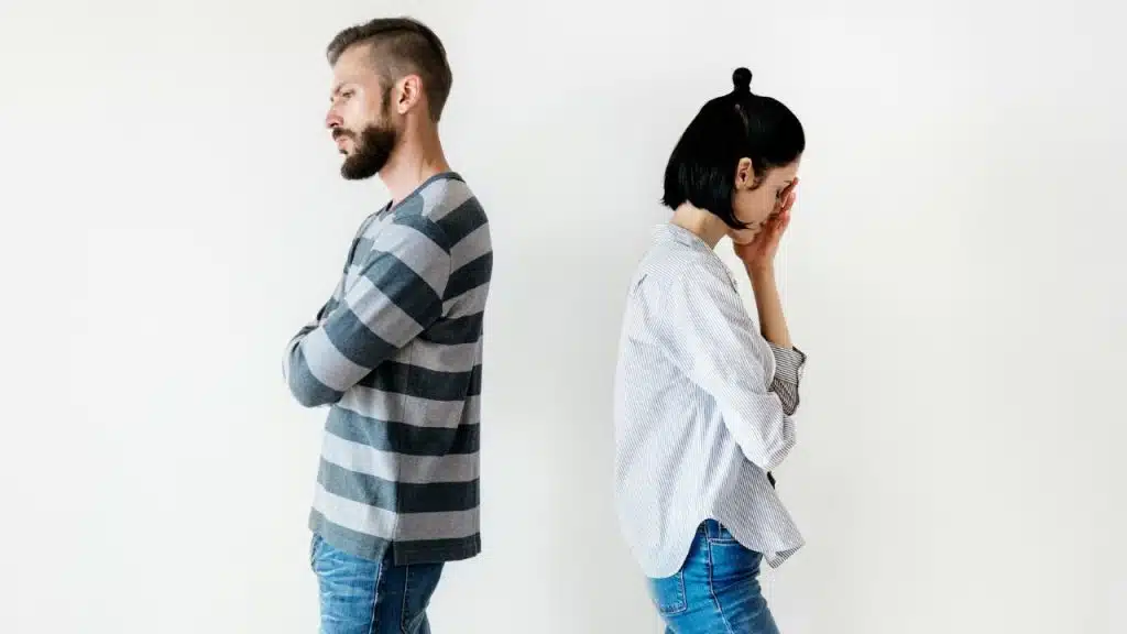 A man and woman stand back-to-back against a white wall; the woman covers her face.