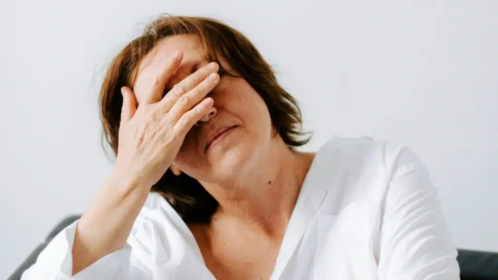 Older woman with brown hair and white shirt covering her face with one hand.