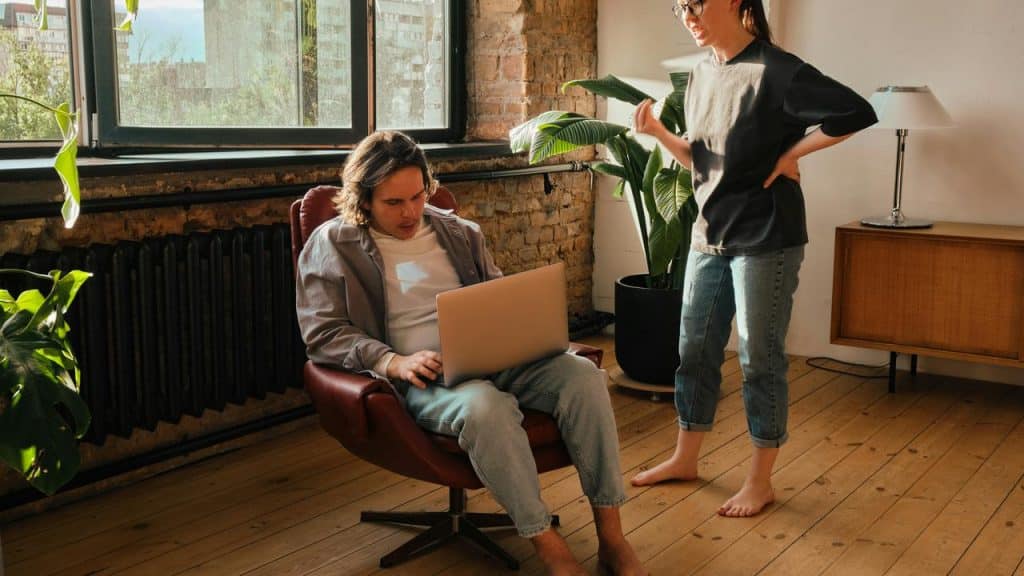 A man sitting on a chair using a laptop while a woman stands nearby talking to him.
