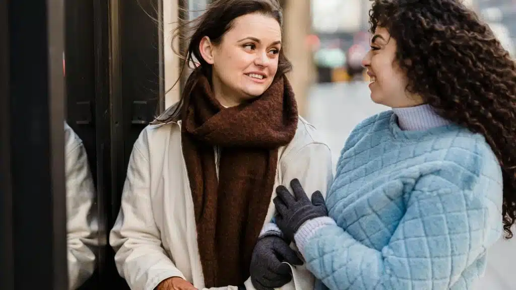 A pair of women standing outside and smiling while talking to each other.