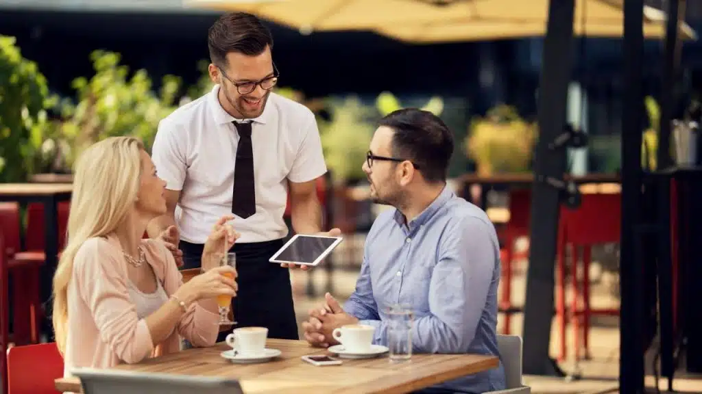 A smiling waiter holds a tablet while talking to a couple seated at an outdoor restaurant table.