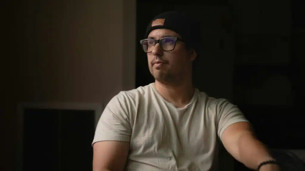 Man in a white t-shirt, black beanie, and glasses looks away in a dark room.