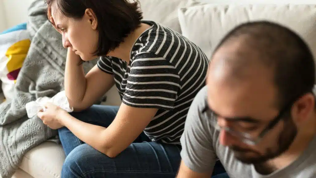 A woman in a black and white striped shirt and jeans sits on a couch looking down sadly, while a blurry man in a gray shirt sits in the foreground.