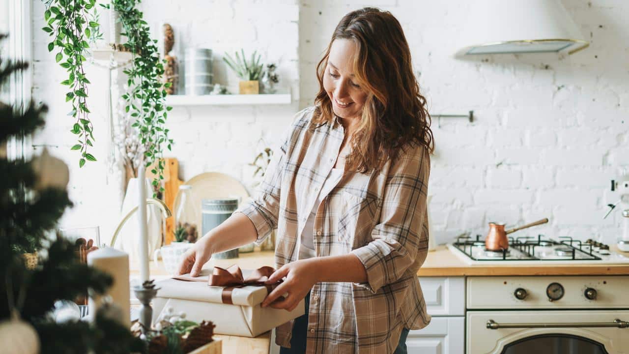A woman in a plaid shirt wrapping a gift in a cozy kitchen.