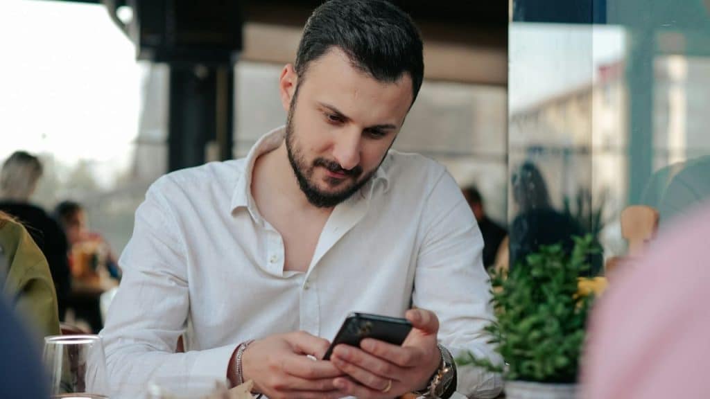 Man in a white shirt looking down at and operating his black cell phone.
