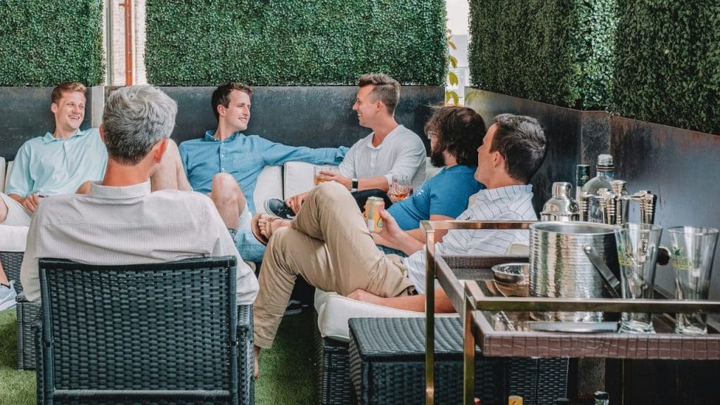Group of men relaxing on outdoor furniture, talking and holding drinks on a patio.