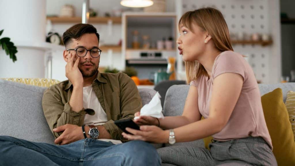 A woman holding a tissue speaks to a man sitting beside her on a couch.